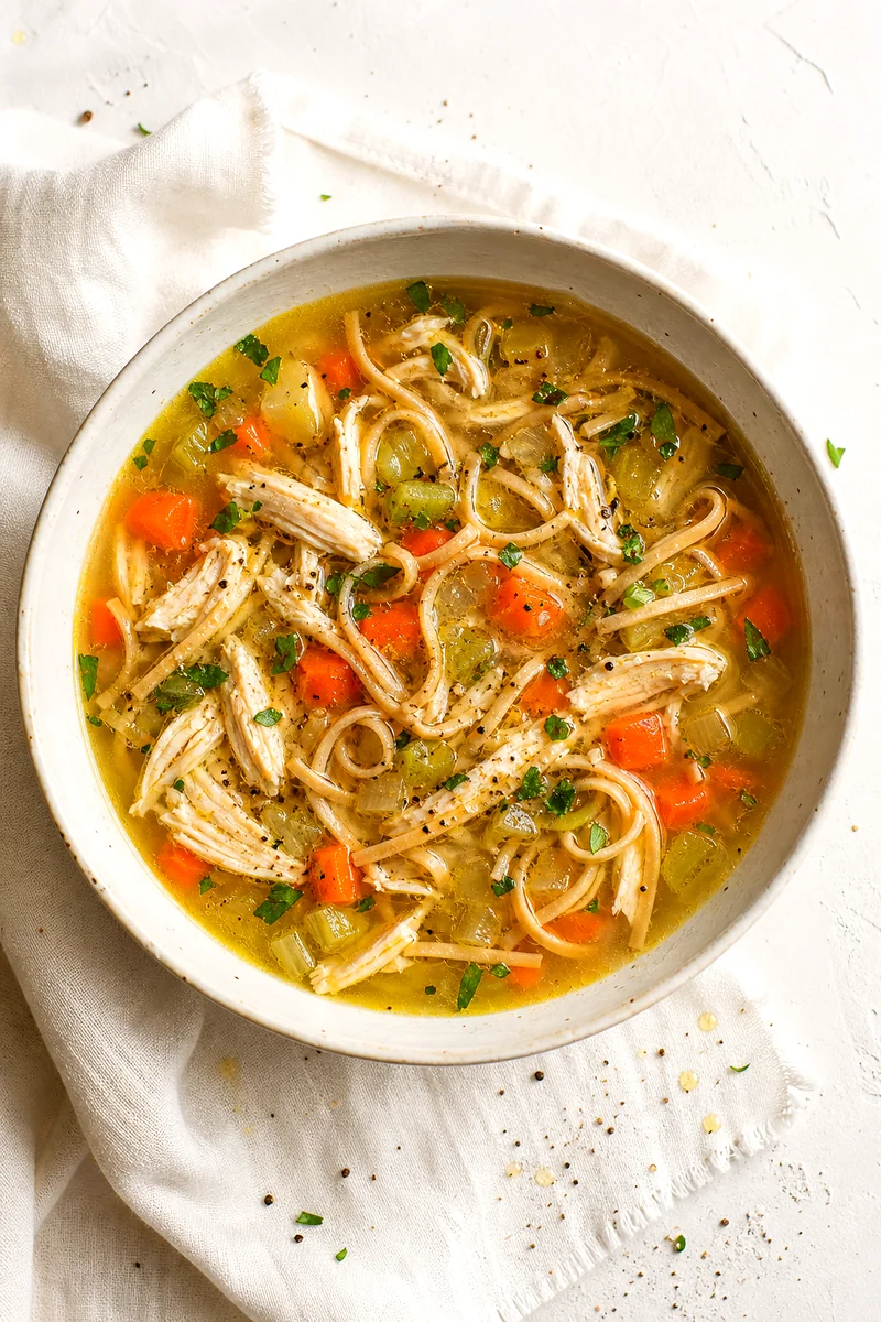 Top down bowl of chicken noodle soup with shredded chicken, noodles, vegetables, parsley, and golden broth