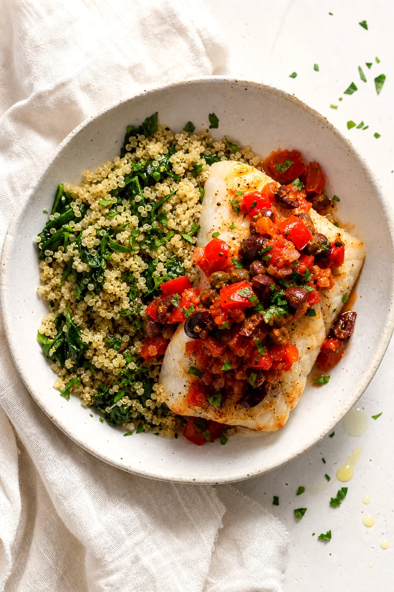 Top down view of baked cod with tomato olive relish and herbed quinoa in a white bowl