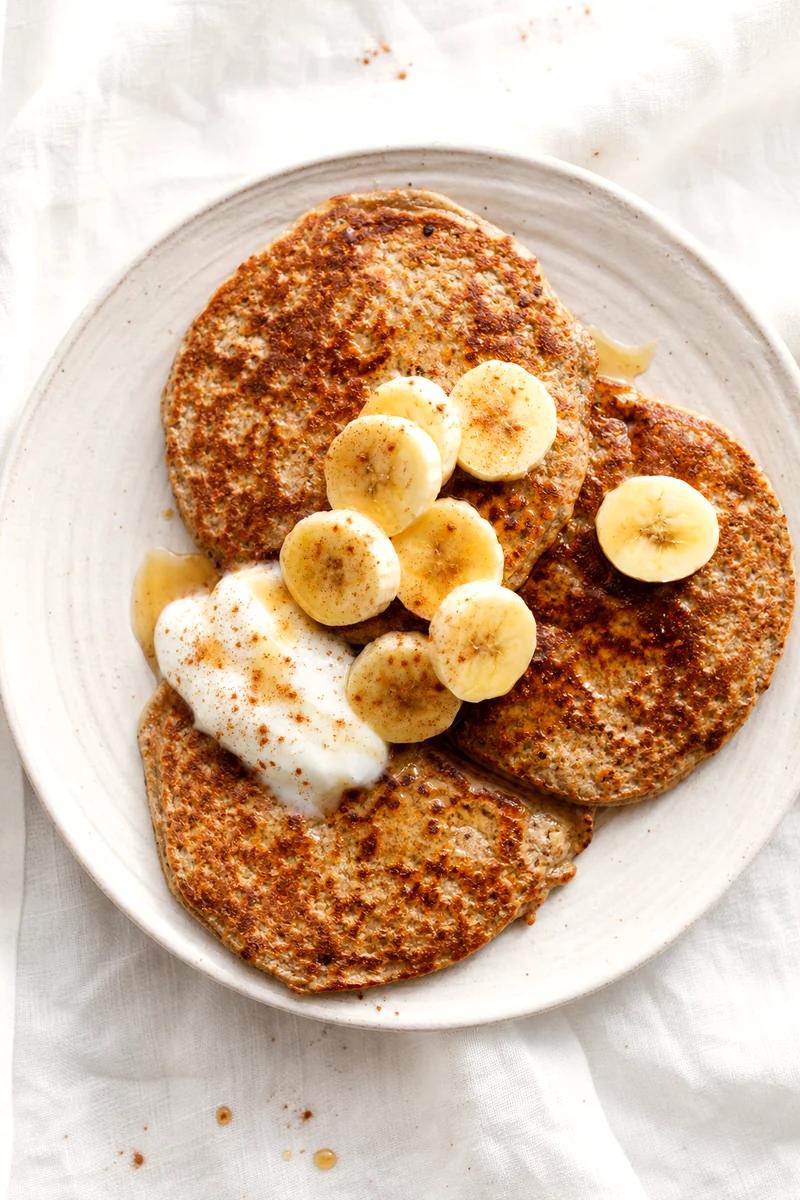 Banana flaxseed pancakes with yogurt, banana slices, cinnamon, and maple syrup on a white plate