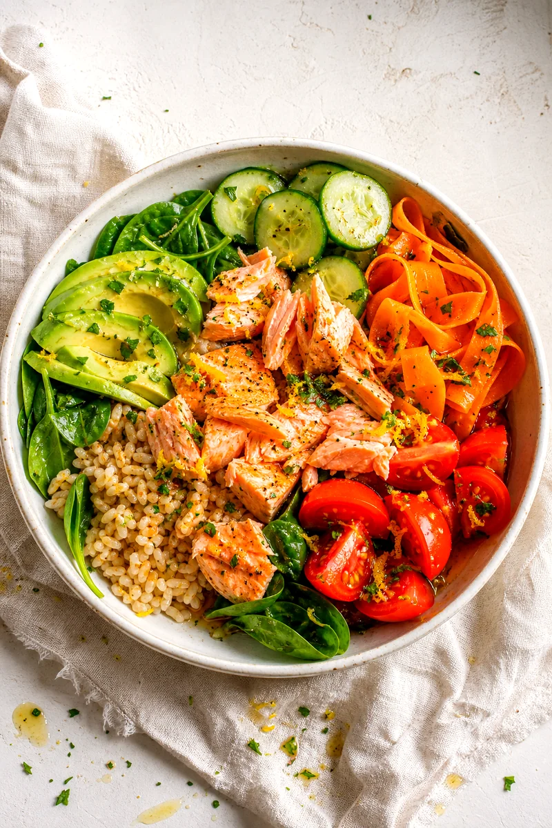 Top view of salmon, brown rice, avocado, cucumber, carrots, spinach, and tomatoes in a white bowl