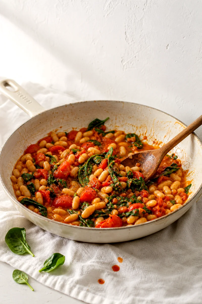 Cannellini beans, tomatoes, spinach, and garlic warming in a skillet with a wooden spoon