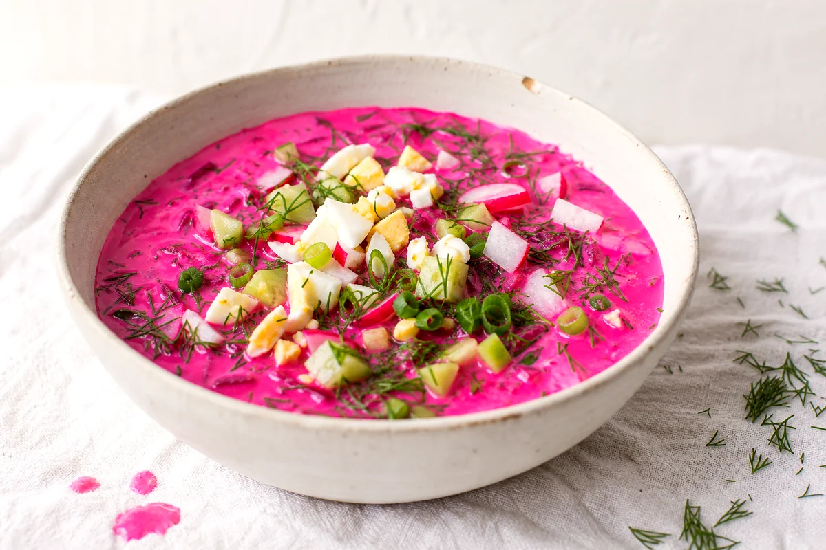 Pink beet and kefir soup topped with chopped egg, dill, cucumber, and radishes in a white ceramic bowl