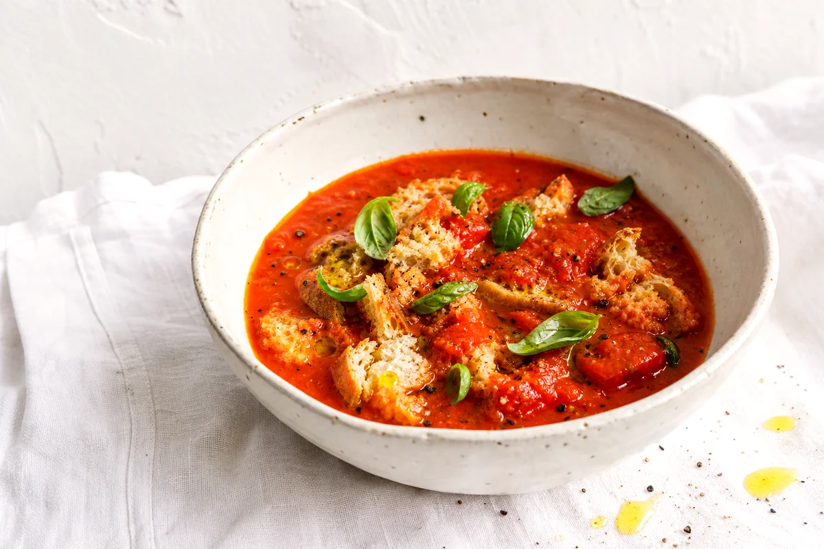 Thick Tuscan tomato soup with torn bread, basil, black pepper, and olive oil in a white ceramic bowl