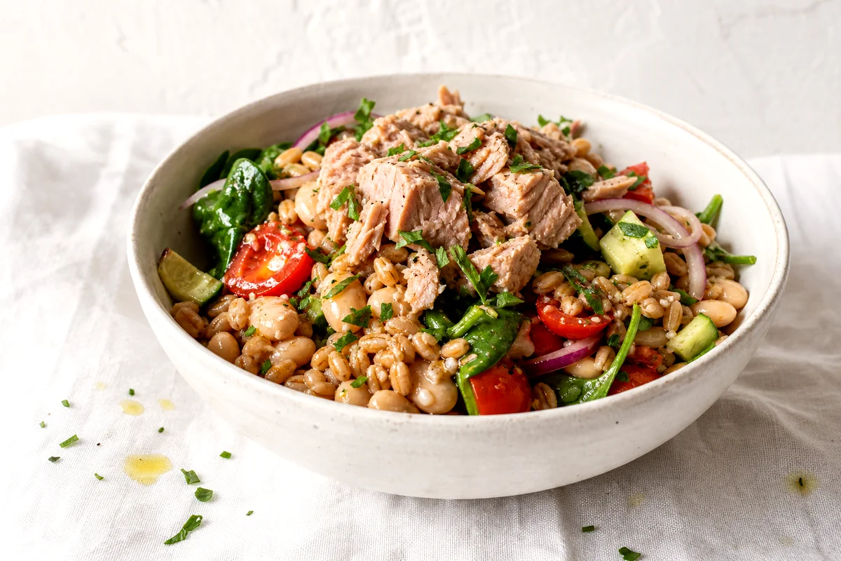 White ceramic bowl filled with tuna, white beans, farro, cucumber, tomatoes, spinach, and parsley